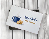 Personalised biscuit tin with the lid resting on the open tin on white wooden boards, watercolour enamel mug and biscuits design