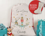 A white drawstring bag with a Christmas-themed design, featuring a nutcracker, a girl, and stars. The bag is labeled "Special Delivery For Charlotte" and is accompanied by a matching stocking. The bag is placed next to a stuffed reindeer and a blue gift box, suggesting it is a Christmas stocking.