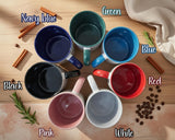 A collection of seven colorful mugs arranged in a circular pattern on a wooden surface, with cinnamon sticks and coffee beans scattered around.