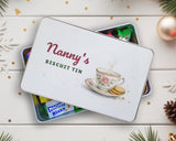 Personalised biscuit tin with "Nanny's Biscuit Tin" lid resting on a tin of sweets, with baubles and pine cones